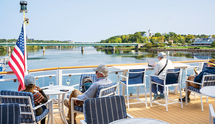A couple sits at a table beside an American flag on an American Cruise Lines Coastal Cat's sun deck looking out at the New England Coast, while a man sits to the right of them typing on his computer.