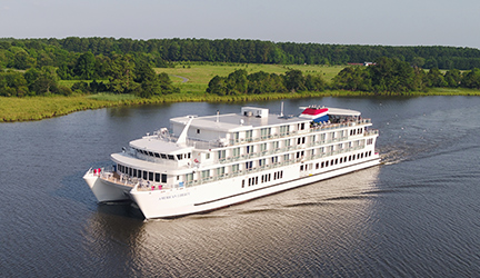 American Cruise Lines' Coastal Cat small cruise ship, American Liberty, sails along a river featuring a beautiful nature landscape.