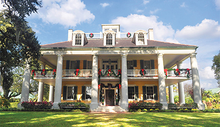 A historic plantation in Louisiana, Houmas House Plantation has two story pillars on all sides, grand porches and is set amongst beautiful grounds and gardens with a red brick pathway. The railings are lined with festive garland and wreaths with red bows