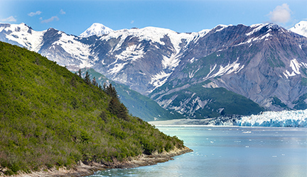 The snow capped mountains in Glacier Bay National Park line the horizon, with a glimpse of an icy blue river in the foreground, beside a hill covered in greenery. 