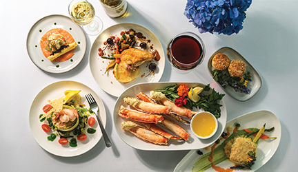 Five plates filled with perfectly-presented cuisine representing the Columbia and Snake Rivers region. The plates include king crab legs, roasted chicken, smoked salmon, a dish of biscuits, a seafood salad, and halibut. There are two glasses of wine beside the plates, all on top of a white surface.