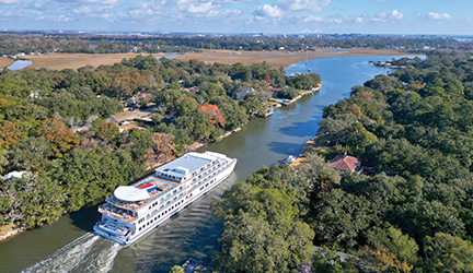 An aerial view of American Liberty, an American Cruise Lines Coastal Cat small cruise ship sailing through Elliot Cut towards Charleston. Each side of the waterway is filled with thick trees and a few buildings throughout. 