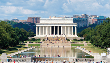 The Lincoln Memorial, a large white building with tall columns around the front entrance with there are several wide stairs leading up to it. There are countless people on the stairs and through the columns. In front of the memorial is the Lincoln Memorial Reflecting Pool, a large, still pool of water surrounded by a concrete pathway with people walking on it. 