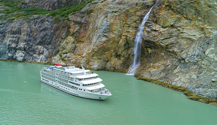 The American Constellation Coastal Cruise Ship sailing through the light green colored passage of Tracy Arm Fjord, surrounded by a cliff with a narrow waterfall.