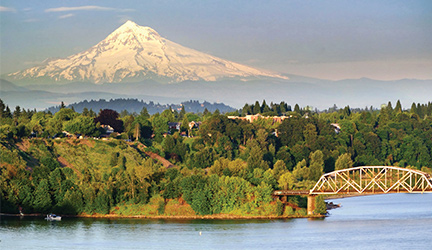 An aerial view of the Portland cityscape, with the Columbia river in the foreground, and a large bridge with a white arch on the top. The cliff beside the river is covered with greenery, and there is a tall, snow-covered mountain in the far distance.