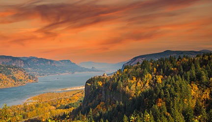 An aerial view of the Columbia River Gorge, featuring a distant view of the Vista House, a building that stands alone on the top of a tall, tree-covered cliff. There are large cliffs covered in trees with green and orange colored leaves on each side of the gorge, under a bright orange sunset.