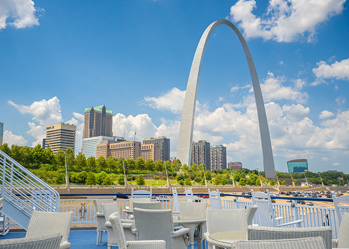 Multiple tables and chairs line the blue American Riverboat deck floor with the view of the St. Louis Gateway Arch and several skyscrapers in the background.