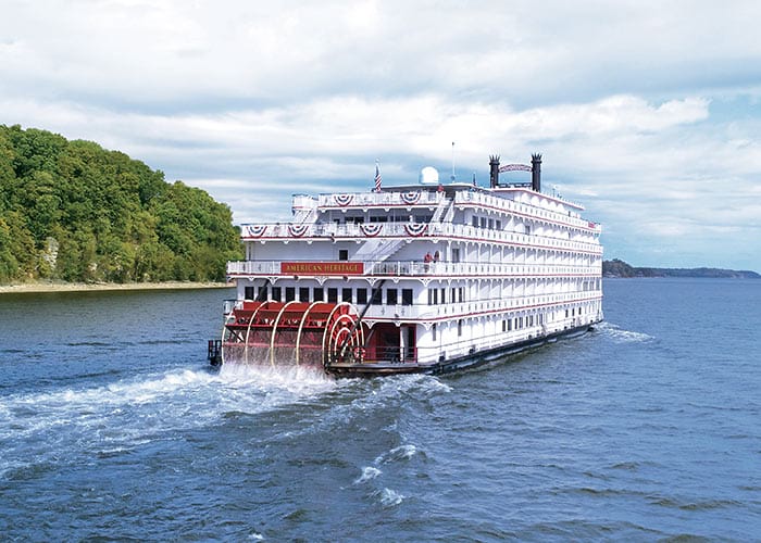 American Melody, an American Riverboat small cruise ship gliding along the Mississippi River, surrounded by lush greenery on the nearby shore.