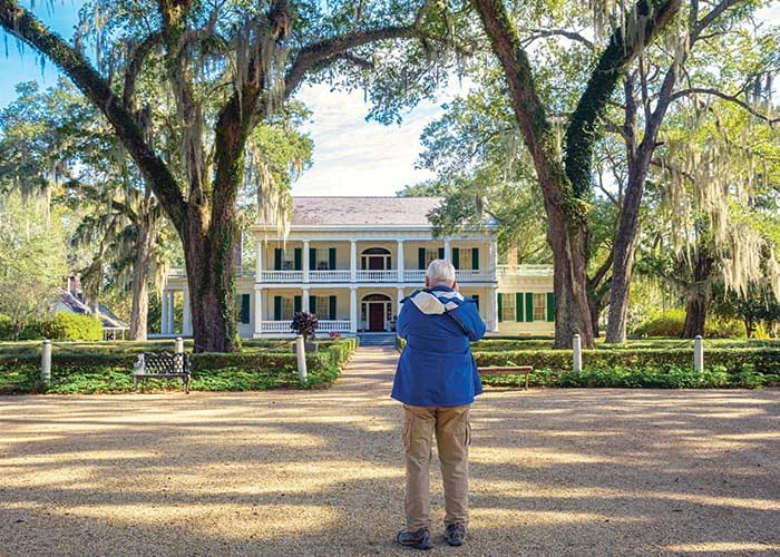 A historic plantation in Louisiana, Oak Alley Plantation, has two story pillars on all sides, grand porches, and is set amongst beautiful grounds and gardens with a red brick pathway with large oak trees on each side leading to the front door. 