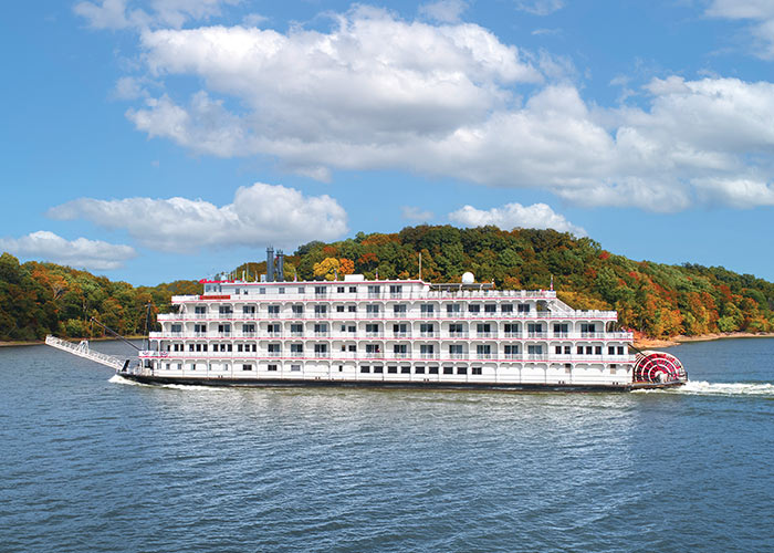 American Melody, an American Riverboat small cruise ship gliding along the Mississippi River, surrounded by lush greenery on the nearby shore.