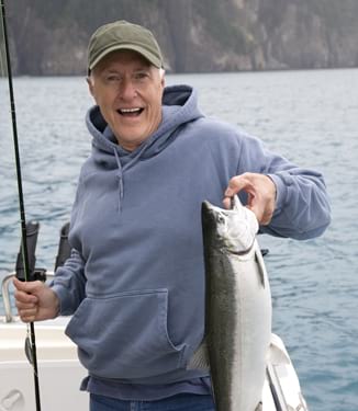 A man smiling while holding a large fish in his hand and a fishing rod in his other hand aboard a private fishing charter excursion.
