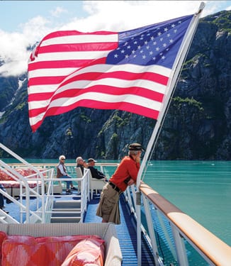 A man leans his arms on the railing of the sun deck on an American Cruise Lines Constellation Class small Cruise Ship next to a large American flag blowing in the wind while sailing along the Inside Passage. There are several people sitting on the sun deck looking at the mountains that line the waters.