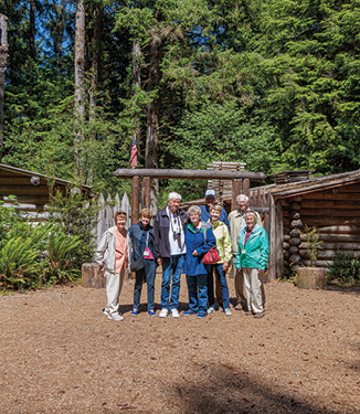 A small group of American Cruise Lines guests smiling for a picture at Fort Clatsop, a historic camp with a wooden gate opening to several other wooden structures in Astoria. There is a flag pole with an American flag flying at the top of it, with tall trees in the background.
