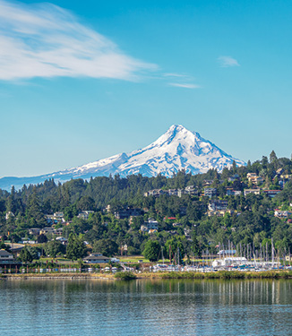 An aerial view of several buildings along a steep hill beside the Columbia River, where there are sailboats docked. The snow-covered Mount Hood peaks over the hill in the distance.