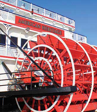 A close up of the back of the American Heritage classic paddlewheeler, showing intricate detail of the red and white paddle wheel and a large red banner with "American Heritage" in gold letters outlined in black on the third deck railing.