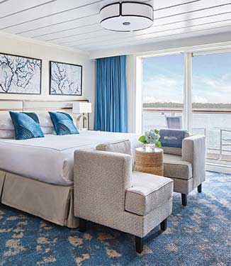 A Veranda Suite on an American Cruise Lines American Riverboat with a white bed with two blue throw pillows, two blue and white paintings above the bed, two accent chairs at the foot of the bed with a small end table that has a plant on top of it, and floor-to-ceiling sliding glass doors that lead to a balcony with scenic views of the Mississippi River.