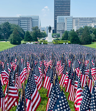 Hundreds of small American flags are placed in rows along the grass in front of a statue of George Washington in Boston's Public Garden. There are several large city buildings and small trees in the distance.