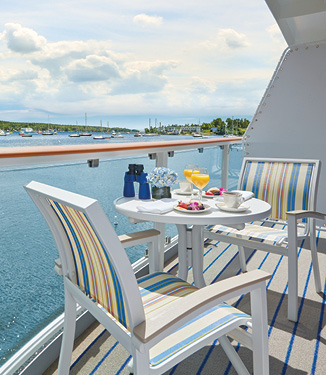 On an American Cruise Lines Coastal Cat private balcony, two blue and yellow striped chairs surround a round, white table with a two plates of food and yellow drinks, binoculars, and a pot with blue flowers. Outside the glass railing is the Intracoastal Waterway.