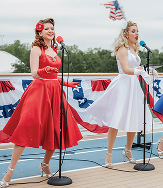 The Victory Belles performing on board for American Cruise Lines passengers. One woman is wearing a red dress with a red flower in her hair, and the other is wearing a white dress with a white flower in her hair.