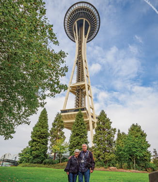 A couple smiling for a photograph while standing in front of the Seattle Space Needle, a tall, narrow structure with a larger circular platform on top.