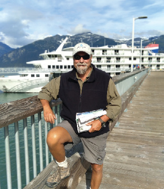 An American Cruise Lines expert guide resting his arm on the railing of a boardwalk over Puget Sound while smiling for a picture with a Constellation Class Coastal Ship docked in the distance.