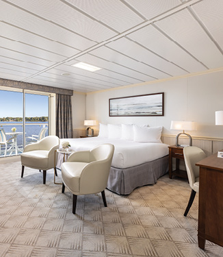 An American Cruise Lines Constellation Class Sky Suite with a white bed with white pillows with a painting on the wall above, two cream colored accent chairs at the foot of the bed, end tables with a lamp at either side of the bed, and floor-to-ceiling sliding glass doors that lead to a private balcony with two chairs and a table overlooking Puget Sound.