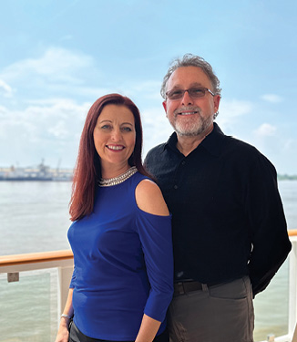Two American Cruise Lines entertainers smiling for a photograph aboard a small cruise ship  with views of the intracoastal waterway in the distance. The woman is wearing a blue blouse and the man is wearing a black button up.