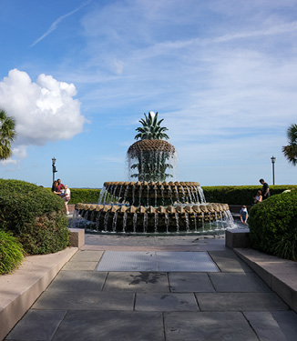 The Pineapple Fountain, a large fountain with water flowing down from layers that looks like a pineapple split in three sections, with the crown as the top layer in Charleston Waterfront Park. Perfectly trimmed bushes surround the circular pool below the fountain and the stone path leading up to it.