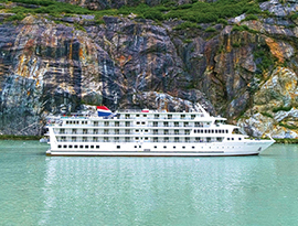 The Constellation Coastal Cruise Ship sailing through the light green colored passage of Tracy Arm Fjord, surrounded by a cliff with bright gold and white streaks down the side.