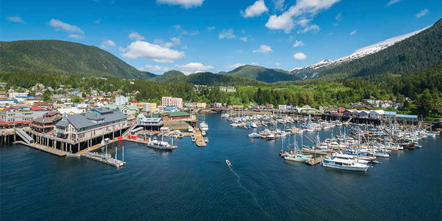 An aerial view of the town of Ketchikan, with several boats docked on the water and the green mountains of Tongass National Forest in the background. 