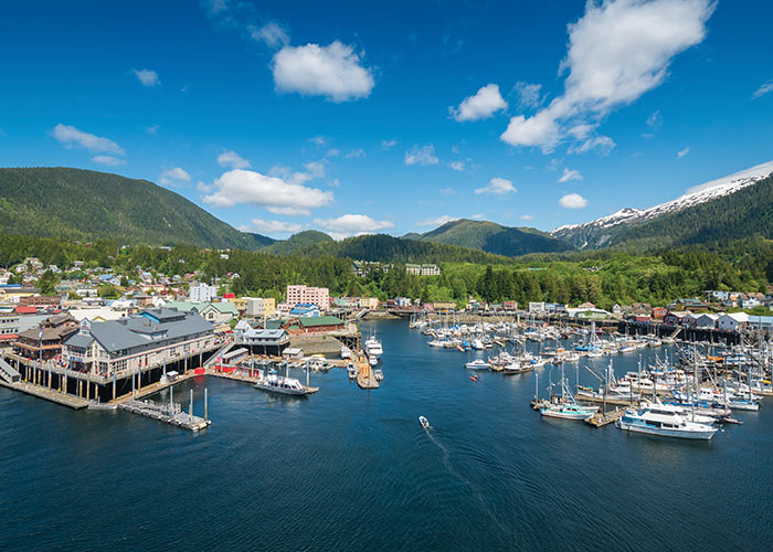 An aerial view of the town of Ketchikan, with several boats docked on the water and the green mountains of Tongass National Forest in the background. 