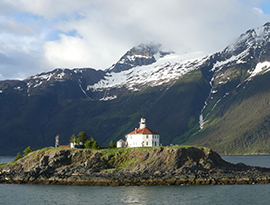 Eldred Rock Lighthouse, a small white building with a brown roof, sits on an island in the middle of the dark blue waters of Lynn Canal in Haines, with the snow-capped mountains in the back.