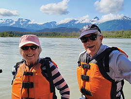 A male and female wearing orange and black life vests, black sunglasses, and baseball caps smiling at the camera with the Inland Passage and expansive mountains in the background.