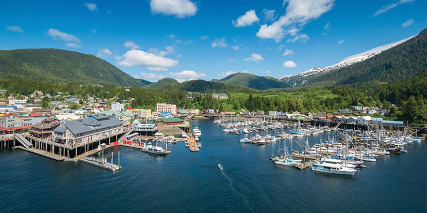 An aerial view of the town of Ketchikan, with several boats docked on the water and the green mountains of Tongass National Forest in the background. 