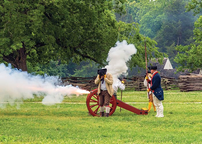 A red-wheeled cannon firing with smoke and a flash of flames shooting from the front and top. Four men dressed in colonial clothing stand beside the cannon, with the one closest to the blast covering his ears. There are thick trees and a log fence in the background.