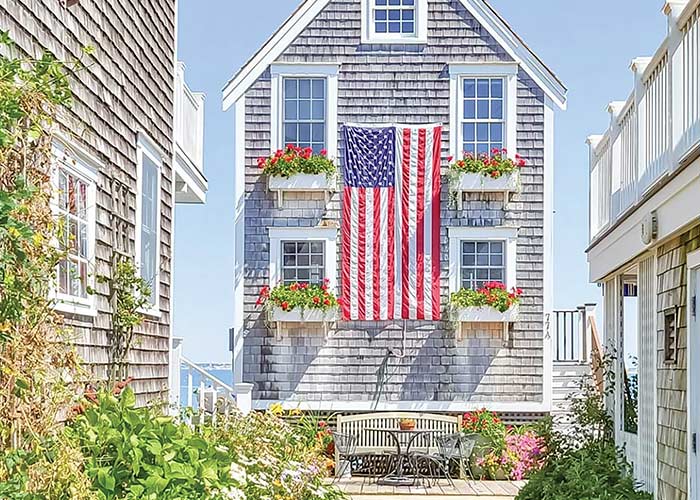 A tall and narrow grey house in Provincetown with four white windows, each with a flower box containing pink flowers, and a large American flag hanging on its side in the center. There is a house very close to each side, flowers and greenery alongside the houses, and a bench in front of the house.