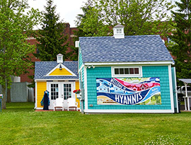 Three small, colorful buildings, the Bismore Park Shanties in Hyannis. The building on the left is olive green and has a red sign on the side reading "Artists". The building on the right is bright blue with a banner on the side reading "Welcome to Hyannis" with different tiers of colors and buildings on it. The building in the middle is dark yellow and has two white chairs in front of it. 