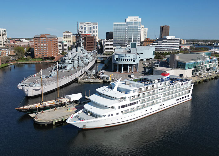 An American Cruise Lines small cruise ship docked in Norfolk, with an aerial view of the cityscape. To the left of the cruise ship, the large Battleship Wisconsin is docked.