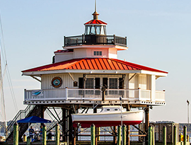 Choptank River Lighthouse in Cambridge. The lighthouse has a red roof over the white balcony, and a small white and red boat beneath the balcony above the water.