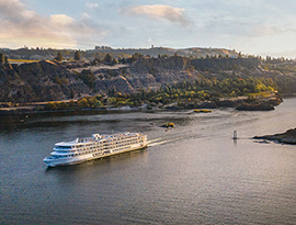 An aerial view of an American Riverboat cruising through the Columbia River Gorge, where there is a small island formation in the middle of the water. Alongside the water, there are several cliffs, and mountains in the distance.