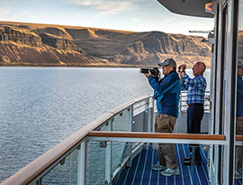 Two American Cruise Lines cruise guests taking photos of the scenic desert canyon mountain views from the deck of an American Riverboat. One of the guests is taking a photo with a large camera, while the other is using his phone camera.