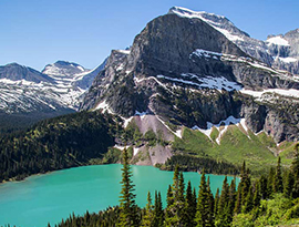 Iceberg Lake, a blue/green pool of water surrounded by snow-capped mountains and tall trees in Glacier National Park.