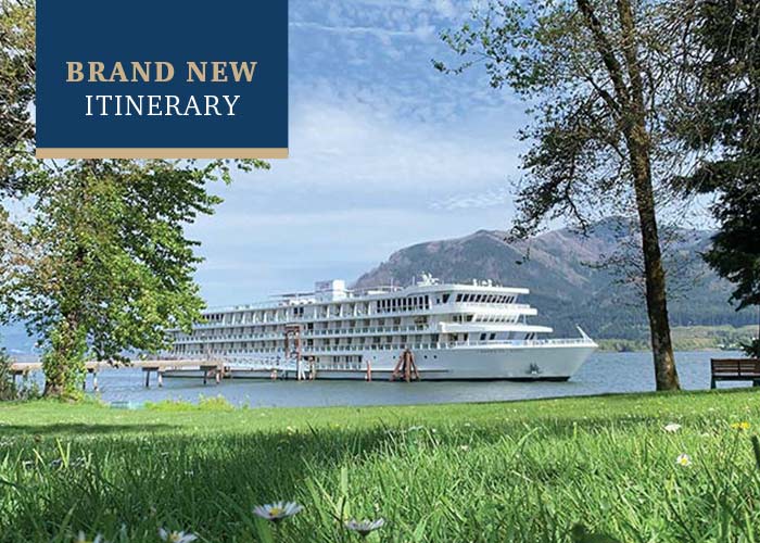 American Song docked along the Columbia River in Richland, where small white flowers peak out from the tall grass. There are trees along the shoreline, a park bench, and mountains in across the river in the distance. In the top left corner, there is a text overlay on a blue box that reads "Brand New Itinerary".