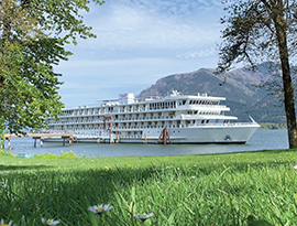 American Song docked along the Columbia River in Richland, where small white flowers peak out from the tall grass. There are trees along the shoreline, a park bench, and mountains in across the river in the distance.