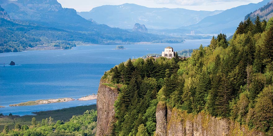 A white building, the Vista House, stands alone on the top of a tall, tree-covered cliff. The Columbia River Gorge with cliffs on the opposite side are in the distance.