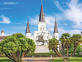 The St. Louis Cathedral with three spires stands in Jackson Square in New Orleans, while a statue of a rider on a horse is centrally placed in a manicured park with trees and bright greenery.