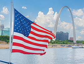 An American flag on a pole connected to the blue and white railing of an American Riverboat blows in the wind along the Mississippi River and the St. Louis Gateway Arch in the distance.