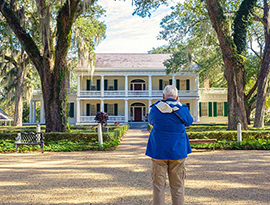 A man wearing a distinct blue jacket stands in front of the historic St. Francisville landmark, the Rosedown, a yellow house with bright green shutters and two-story white pillars with a brick pathway surrounded by perfectly trimmed bushes and large trees leading to the red door. 