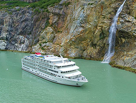 The American Constellation Coastal Cruise Ship sailing through the light green colored passage of Tracy Arm Fjord, surrounded by a cliff with a narrow waterfall.