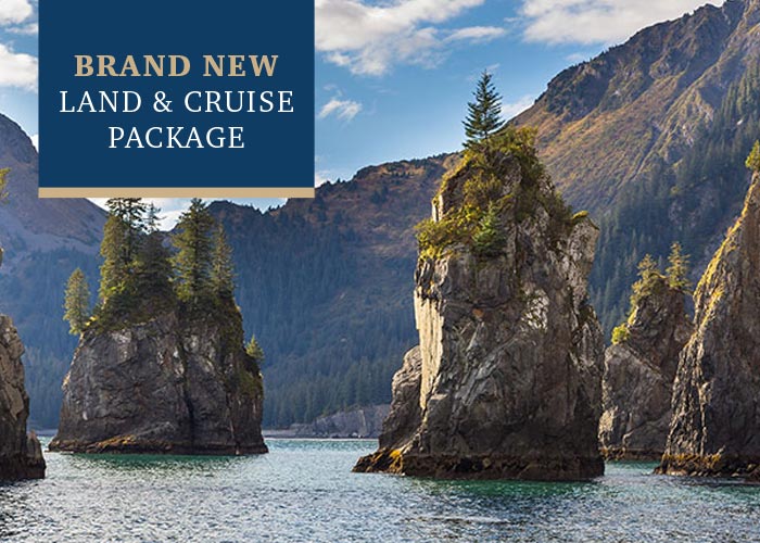 Mountains stand tall around the waters of Kenai Fjords National Park. There are a few tall and narrow  mountain-like structures that are surrounded by the water. In the top left corner, there is a text overlay on a blue box that reads "Brand New Land & Cruise Package".